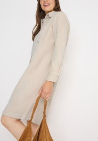 Beige, randig skjortklänning med långa ärmar, bröstficka och en avslappnad passform. Modellen håller en brun mockaväska.