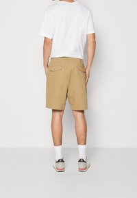 Shorts khaki con due tasche posteriori, indossati con una t-shirt bianca. Calze bianche visibili e scarpe da ginnastica grigie con accenti rossi.