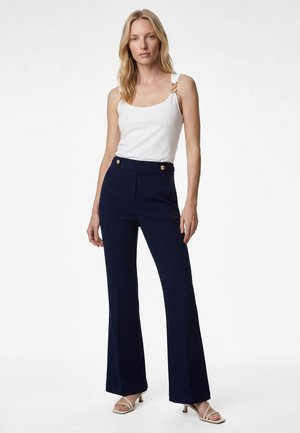 Mujer rubia con una blusa blanca sin mangas con anillos dorados en las tiras, pantalones azul marino de cintura alta con botones dorados y sandalias beige de tacón.