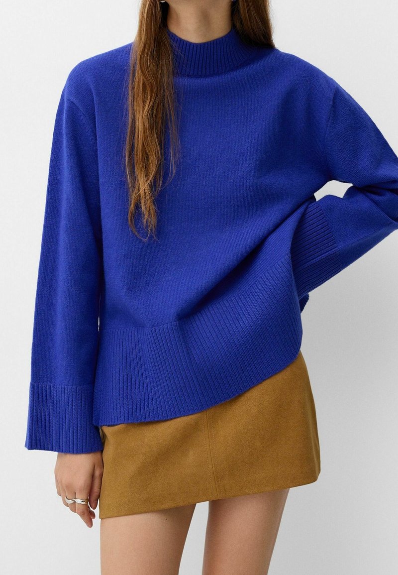 Femme portant un pull côtelé bleu royal et une mini-jupe en daim beige, se tenant debout avec une main sur la hanche, sur fond blanc.