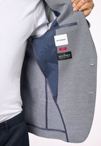 Interno di un blazer grigio con fodera blu, etichette e tasche. Il materiale presenta un motivo texturizzato. Design slim fit, adatto per abbigliamento formale.