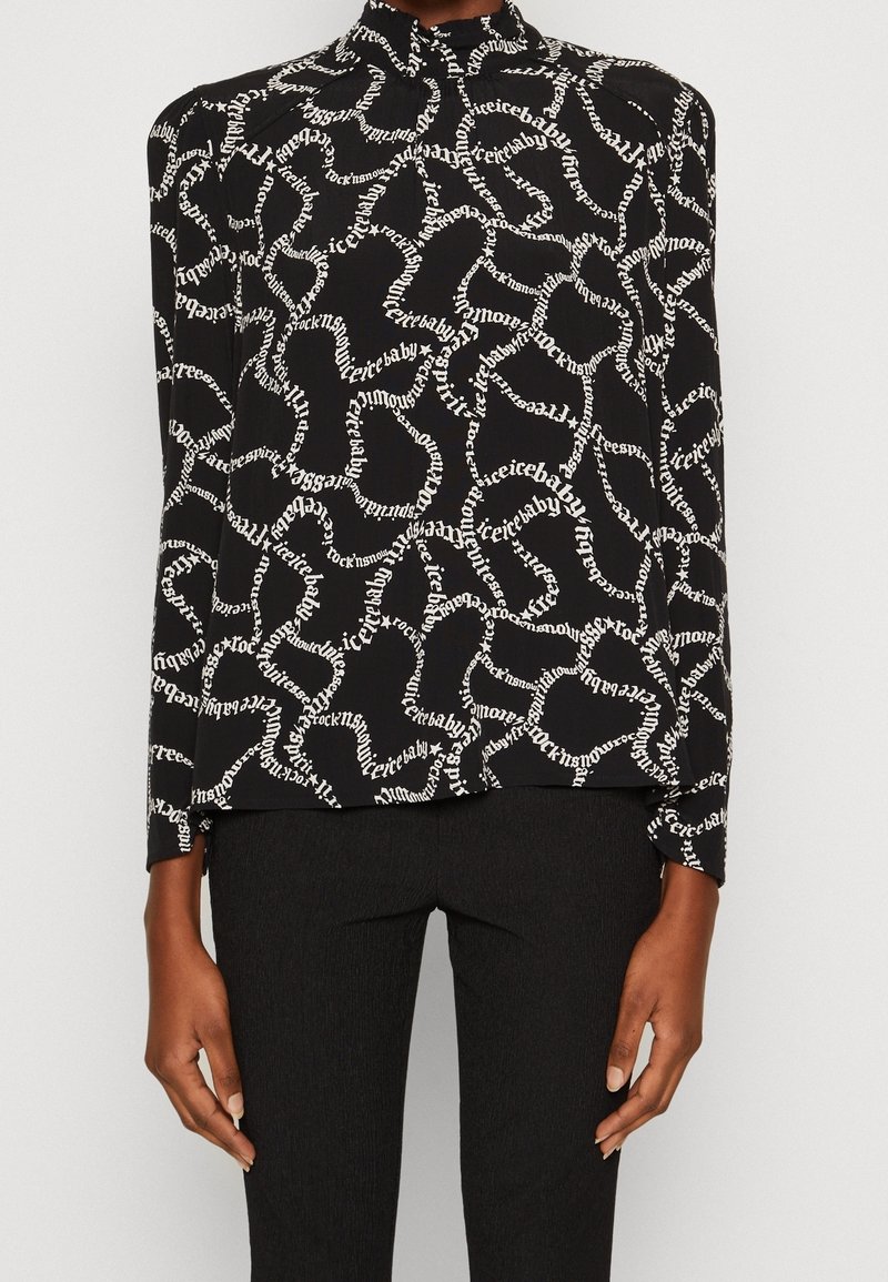 Haut noir à manches longues avec motifs de mots ondulés blancs et col montant, porté avec un pantalon noir ajusté, bras détendus sur les côtés.
