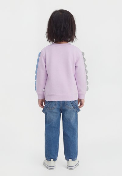 Παιδί με σκούρα μαλλιά μέχρι τους ώμους φοράει ένα μωβ φούτερ και φαρδιά μπλε τζιν, στέκεται γυρισμένο προς το λευκό φόντο.