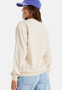 Crèmekleurige sweatshirt met een relaxed fit, geribbelde manchetten en een geribde onderkant. Gecombineerd met lichtblauwe jeans en een blauwe pet. Minimalistisch ontwerp.