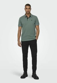 Poloshirt aus grünem Baumwollstoff, mit einem schwarzen Kragen und weißen Akzenten. Kombiniert mit schwarzen Hosen und schwarzen Schuhen, in aufrecht stehender Haltung.
