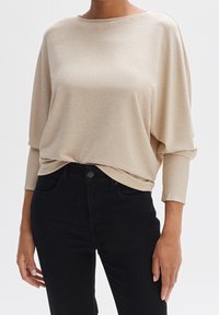 Haut beige à manches longues côtelé avec un large col, coupe décontractée et texture subtile, associé à un jean noir ajusté.