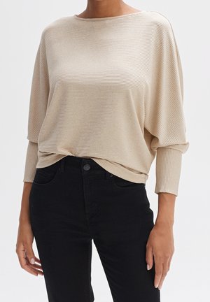 Beige ribbed long-sleeve top with a wide neckline, relaxed fit, and subtle texture, paired with black fitted jeans.