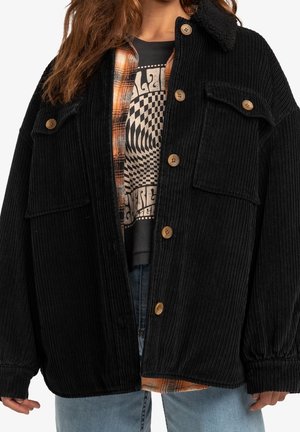 Personne portant une veste boutonnée en velours côtelé noir avec poches à rabat par-dessus une chemise à carreaux et un t-shirt graphique, associée à un jean bleu.