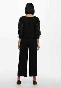 Vrouw met kort zwart haar, staand met de rug naar de kijker, gekleed in een zwart kanten top met pofmouwen, zwarte wijde broekspijpen en zwarte hoge hakken.