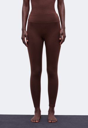 Person står barfodet iført mørkebrune tætsiddende leggings og matchende top mod en ensfarvet lys baggrund.