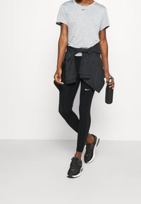 Grå Nike-t-shirt, svarta leggings med logo, svart ytterkläder knutna runt midjan, håller en vattenflaska, bär svarta träningsskor.