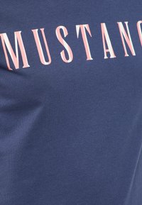 Himmelblaues Baumwoll-T-Shirt mit großem "MUSTANG"-Text in Korallenrot und Weiß, mit einer glatten Textur und tailliertem Design.