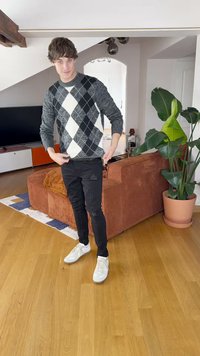 Šedý a černý argyle svetr s bílými diamanty, černé roztrhané kalhoty a bílé tenisky. Dřevěná podlaha a zelená rostlina v pozadí.