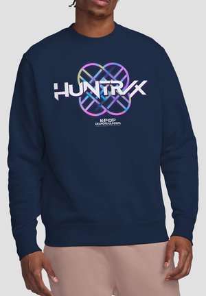 Mann trägt marineblaue Sweatshirt mit buntem "HUNTRVX"-Text und kreisförmiger Grafik über dem Schriftzug "KPOP DEMON HUNTERS" auf der Vorderseite.