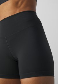 Gros plan sur une personne portant un short de sport noir taille haute, se concentrant sur la taille et la partie supérieure de la cuisse contre un fond uni.