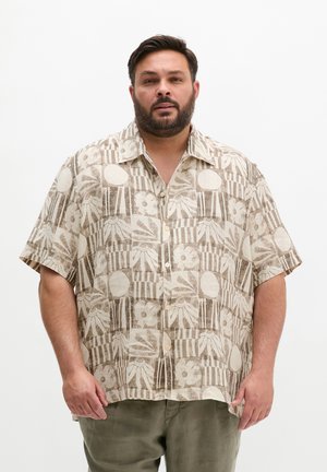 Mężczyzna z brodą, ubrany w beżową koszulę na krótki rękaw z kwiatowymi i geometrycznymi wzorami oraz oliwkowozielone spodnie, stoi na białym tle.