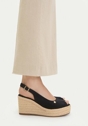 Fod iført beige vide bukser og sorte åbne kile-sandaler med flettet platform og ankelrem med guldbøjle.