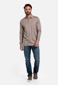 Langarm-Polohemd aus leichtem Beigefarbene Stoff mit Kragen und Drei-Knopf-Leiste, kombiniert mit blauen Jeans und braunen Sneakers.