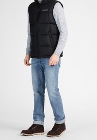 Svart pufferväst med hög krage och dragkedja, bärs över en grå långärmad tröja; matchad med ljusblå jeans och bruna snörkängor.