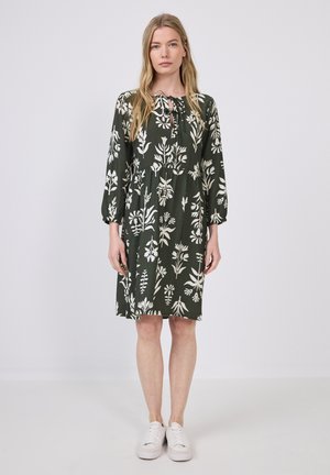 Grünes Kleid mit weißem Blumenmuster, knielang, runder Ausschnitt und langen Ärmeln. Getragen mit weißen Turnschuhen. Glatte Textur.