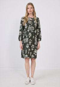 Tmavě zelené šaty s bílými květinovými vzory, s kulatým výstřihem, dlouhými rukávy a nařaseným pasem. Kombinované s bílými teniskami.