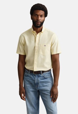 Mężczyzna ubrany w bladożółtą, krótką koszulę na guziki z kieszonką i niebieskie dżinsy, stojący na jednolitym jasnym tle.