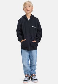 Blond chlapec stojí s rukami v nohaviciach, oblečený v čiernej zipsovej mikine, svetlomodrých džínsach a čierno-oranžových teniskách na bielom pozadí.