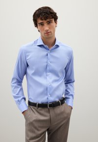 Camicia azzurra chiara con bottoni in cotone, con colletto aperto, maniche lunghe e una texture liscia; indossata con pantaloni beige e una cintura nera.
