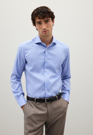Camisa de botones azul claro hecha de algodón, con cuello extendido, mangas largas y una textura suave; usada con pantalones beiges y un cinturón negro.