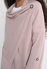 Hellrosa gestrickter Pullover mit locker fallendem Rollkragen, Knopfdetails an der Schulter und samtiger Struktur an den Ärmeln.