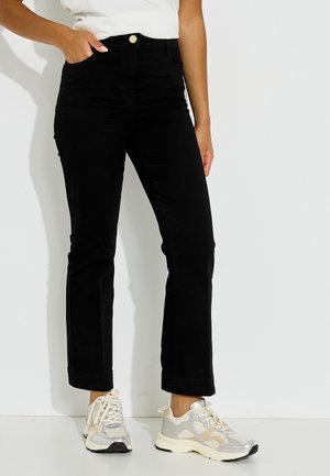 Persona con pantalones negros de pierna recta, camisa blanca y zapatillas plateadas y blancas, de pie con una mano en el bolsillo sobre fondo blanco.