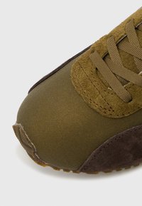 Κοντινό πλάνο από μύτη αθλητικού παπουτσιού σε ελιά και καφέ σουέντ με κορδόνια που ταιριάζουν, σε λευκό φόντο.