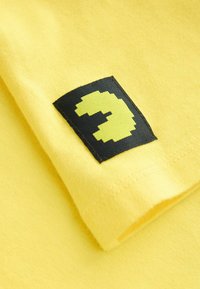 Gul bomulds t-shirt med en foldet design, der prydes af en sort lap med en pixeliseret gul karakter formet som et "3."