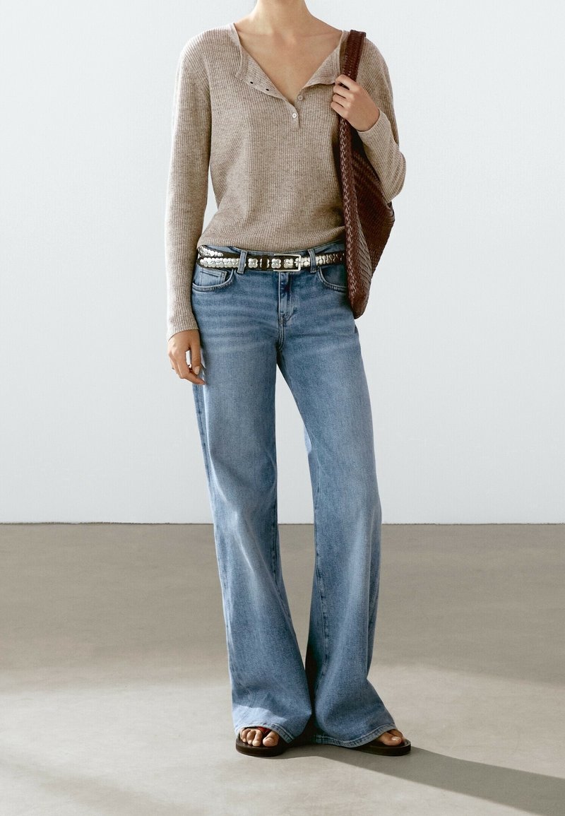 Personne portant un haut beige à manches longues, un jean bleu à jambes larges avec une ceinture cloutée, tenant un sac à bandoulière en tissu marron, et portant des sandales foncées.