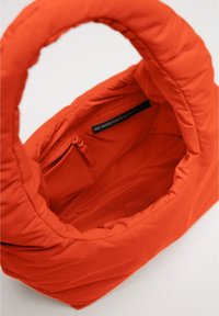 Borsa a tracolla arancione imbottita con una texture morbida, dotata di una tasca con zip interna e una maniglia arrotondata. Il materiale principale è costituito da reti da pesca riciclate.
