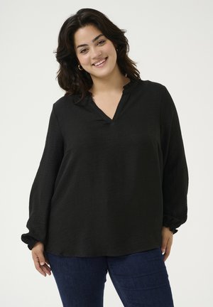 Blouse noire à manches longues avec un col en V et un col à volants, en tissu texturé et avec des poignets élastiqués, associé à un jean bleu foncé.