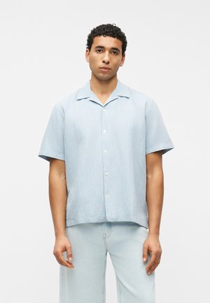 Moški, oblečen v svetlo modro srajco s kratkimi rokavi in belimi navpičnimi črtami ter svetlo modre kavbojke, stoji pred enobarvnim belim ozadjem.