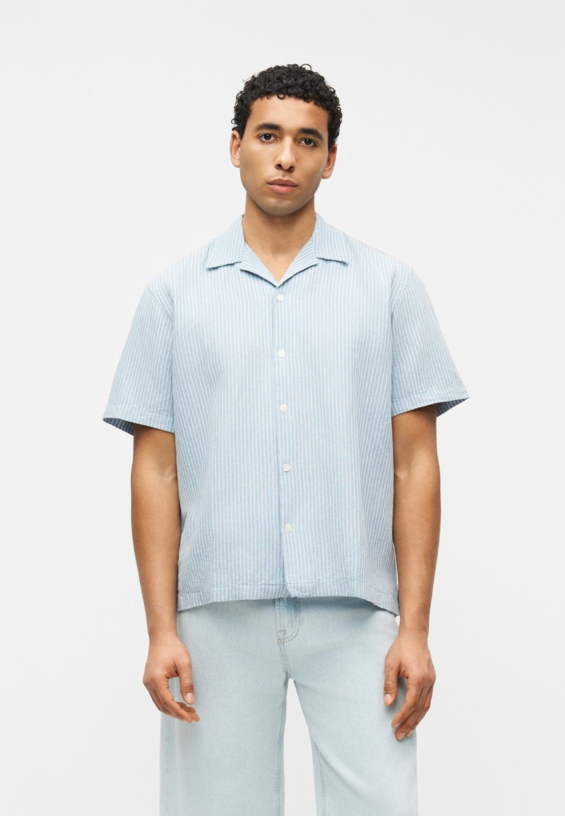 Άνδρας που φοράει ανοιχτό μπλε κοντομάνικο πουκάμισο με λευκές κάθετες ρίγες και ανοιχτό μπλε τζιν, στέκεται μπροστά σε απλό λευκό φόντο.