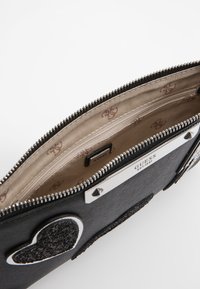 Portefeuille noir avec un extérieur texturé, une quincaillerie argentée et un appliqué en forme de cœur. L'intérieur présente une doublure beige avec imprimé logo.