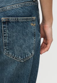 Πίσω τσέπη τζιν με πορτοκαλί ραφές, φορεμένη από άτομο με το δεξί του χέρι χαλαρό στο πλάι.