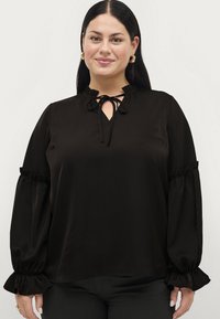 Czarna bluzka z wysokim kołnierzem, zawiązywana na wstążkę, z falbaniastymi rękawami. Wykonana z gładkiego materiału, z delikatnym połyskiem i luźnym krojem.
