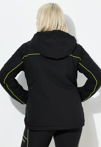 Blondýna viděná zezadu, která má na sobě černou bundu s kapucí se světle žlutým lemováním a černé legíny se světle zeleným prošitím.