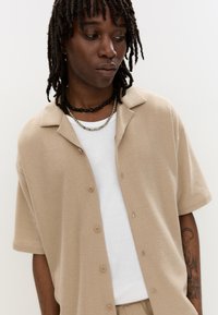 Beige, lyhythihaista, napitettava paitaa, jossa on teksturoitu kangas. Päällä valkoinen, ribattua crew-neck t-paita. Kerrokselliset kaulaketjut viimeistelevät ilmeen.