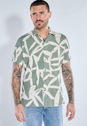 Mann mit kurzen Haaren und Tattoos, der ein kurzärmeliges Hemd mit grün-weißem abstraktem Muster und hellblaue Jeans trägt.