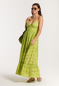 Lime green maxi suknelė su V formos iškirpte, gėlėmis išpjaustytais raštais ir sluoksniuotu apvadu. Dėvima su akiniais nuo saulės ir tekstūruota žalia rankine.