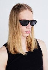Gafas de sol rectangulares negras con lentes oscuros y acabado mate, llevadas por una persona con largo cabello rubio liso, vestida con una camiseta sin mangas negra.
