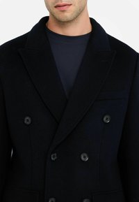 Man in een donkerblauwe dubbelzijdige jas met zwarte knopen over een marineblauwe ronde hals T-shirt, kort boven het onderste gezicht en de schouders.