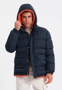 Chaqueta acolchada azul marino con forro de capucha naranja, que cuenta con dos bolsillos frontales, puños elásticos y un cierre de botón a presión.