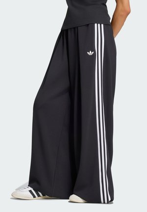Personne portant un pantalon large noir Adidas avec trois bandes blanches sur les côtés et des baskets blanches Adidas avec des bandes noires.