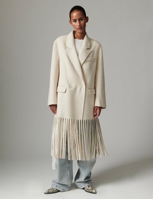 Κρεμ παλτό διπλού πέτου με φουντίτσα στο τελείωμα, κατασκευασμένο από υφήνωμένο μαλλί, με δύο μπροστινές τσέπες, φορεμένο πάνω από φαρδιά τζιν με ανοικτό πλύσιμο.